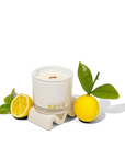 Neve scented white candle with wooden wick surrounded by green leaves and fresh lemon on white surface
