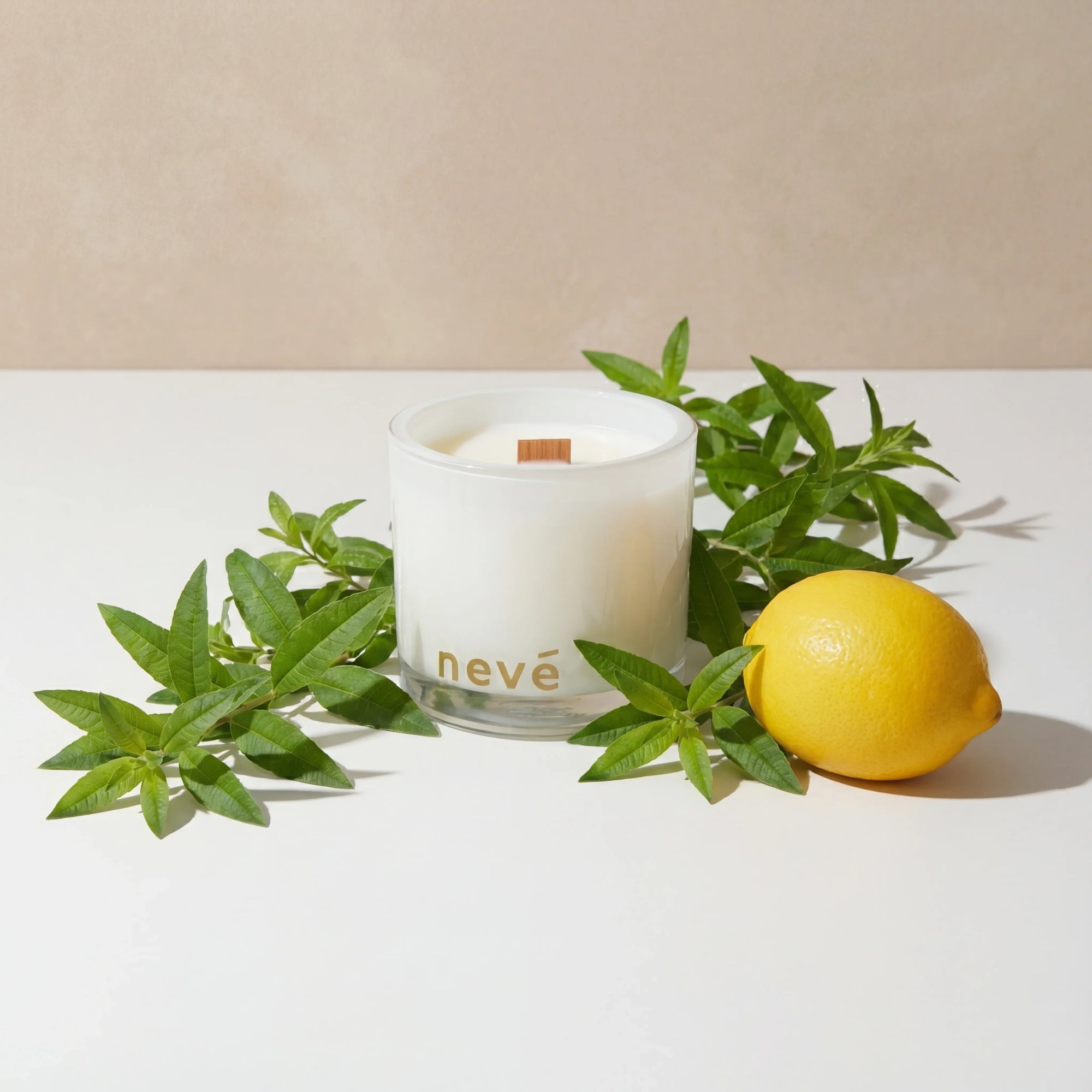 Neve scented white candle with wooden wick surrounded by green leaves and fresh lemon on white surface