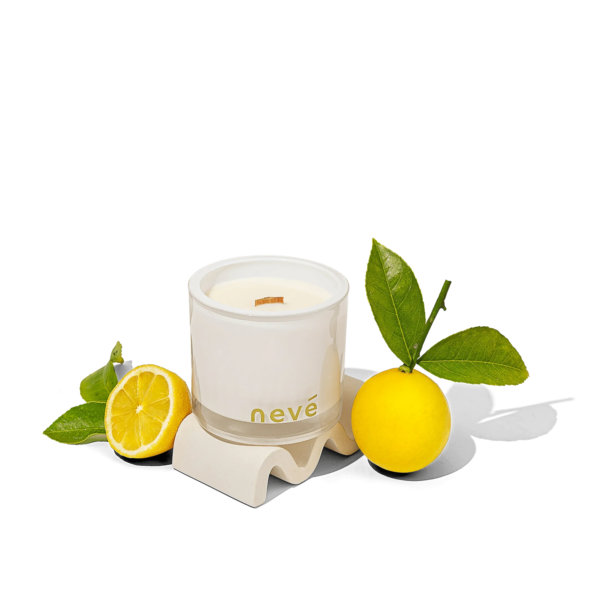 Neve scented white candle with wooden wick surrounded by green leaves and fresh lemon on white surface